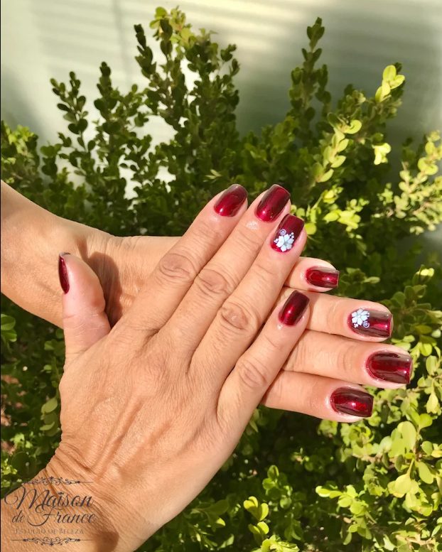 Unhas em bordô metalizado com flor delicada, fotografadas no jardim do salão